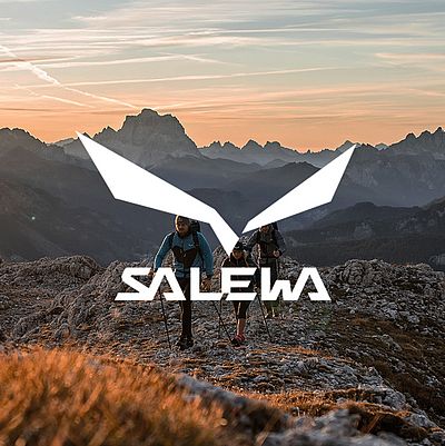 Due uomini e una donna camminano al tramonto su un terreno roccioso. Sull'immagine è riportato il logo Salewa.