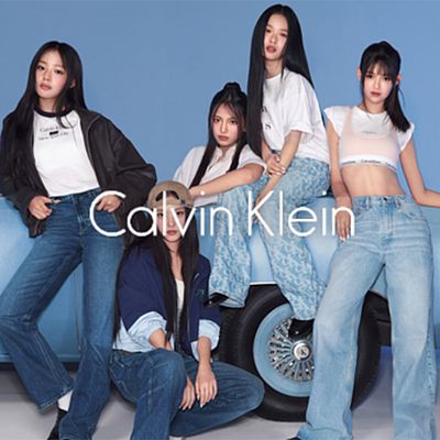 Cinque ragazze vestite con jeans e maglietta sono sedute e appoggiate sul cofano di un'auto. Il logo di Calvin Klein è visibile al centro dell'immagine.