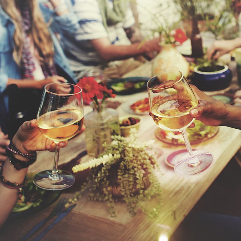 Una tavola imbandita; sopra di essa, due persone brindano con un bicchiere di vino ciascuno.