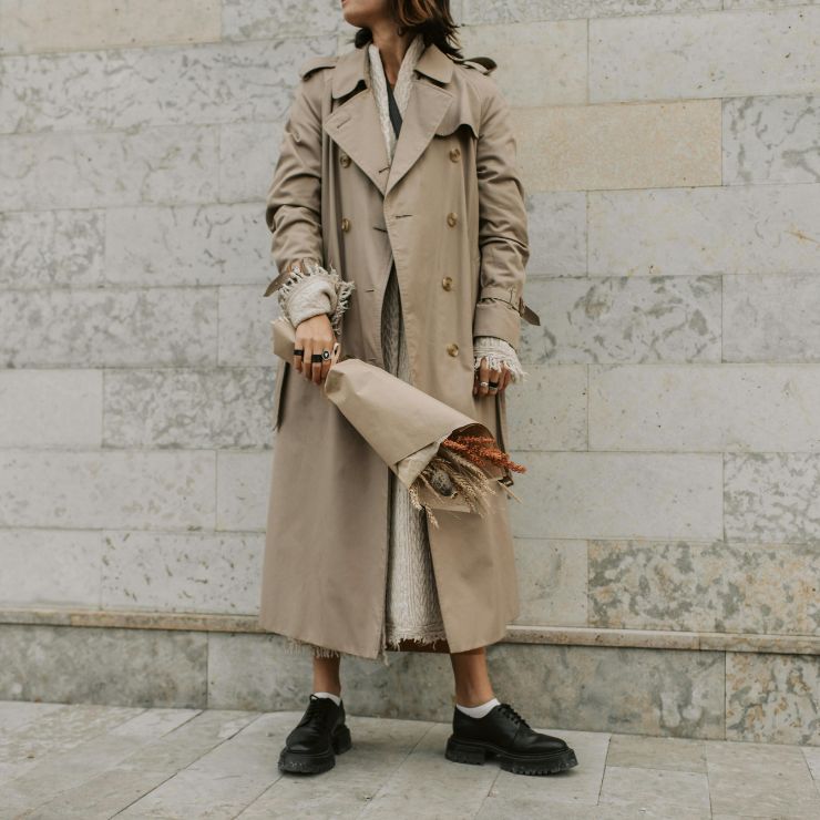 Una donna indossa un trench e tiene un mazzo di fiori nella mano destra.