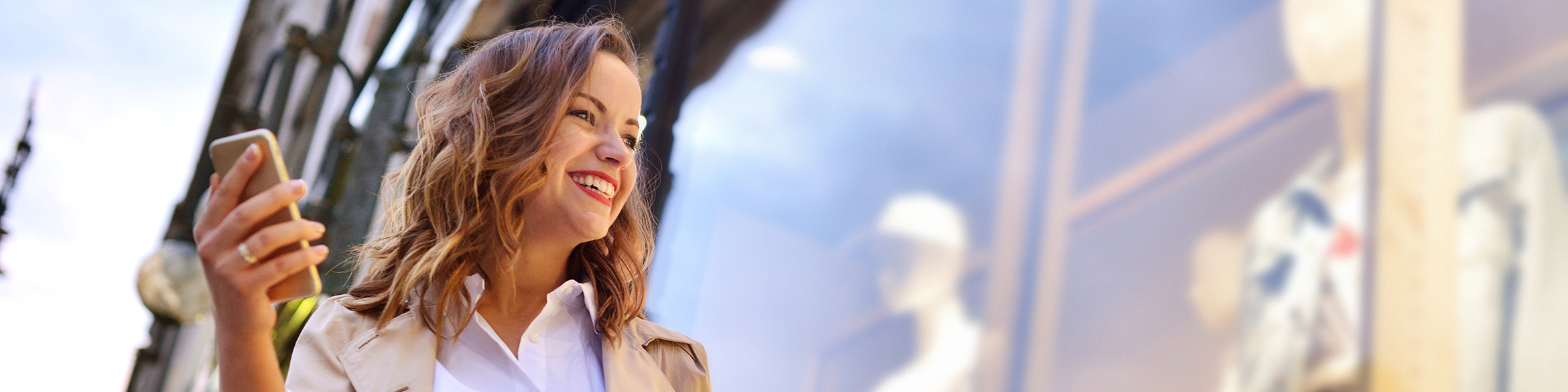 Una donna è in piedi davanti a una vetrina, sorride e tiene un cellulare nella mano destra.