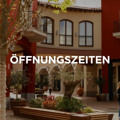 L'immagine mostra una parte del centro commerciale, un passaggio e, sullo sfondo, i negozi fiancheggiati da archi a tutto sesto. Sopra c'è scritto in tedesco: “Orari di apertura”.