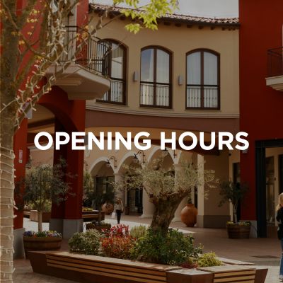 L'immagine mostra una parte del centro commerciale, un passaggio e, sullo sfondo, i negozi fiancheggiati da archi a tutto sesto. Sopra c'è scritto in inglese: “Orari di apertura”.